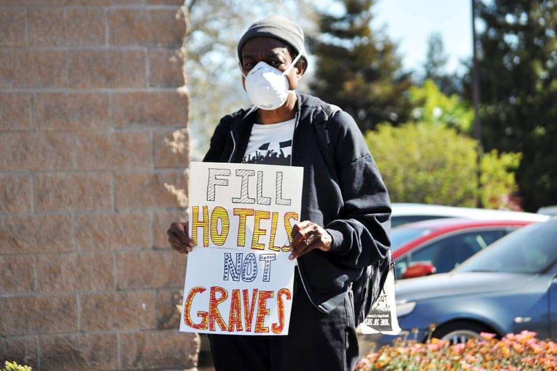 Fill-hotels-not-graves-sign-held-by-Black-organizer-for-East-Oakland-Collective-The-Village, COVID-19 further burdens Oakland’s Black homeless population’s quest to survive, Local News & Views 