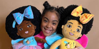 11-year-old doll creator Esi Orijin, holding one doll on the right with large blue bow in her hair with matching dress. Doll on left arm is wearing a yellow bow with a matching dress.Both dolls depict the variations of African American skin color.