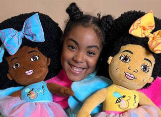 11-year-old doll creator Esi Orijin, holding one doll on the right with large blue bow in her hair with matching dress. Doll on left arm is wearing a yellow bow with a matching dress.Both dolls depict the variations of African American skin color.