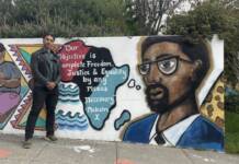 Candidate for governor Dr. Butch Ware joins teachers at Malcolm X Academy in Hunters Point African American male standing with hands clasped in front of him. He is standing in from of Malcolm X mural. The mural is vibrant and the continent of Africa is drawn depicted into three colorful regions. West inked, central in black, and eastern in blue.