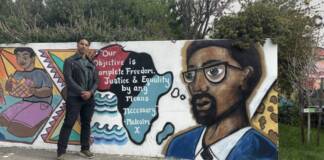 Candidate for governor Dr. Butch Ware joins teachers at Malcolm X Academy in Hunters Point African American male standing with hands clasped in front of him. He is standing in from of Malcolm X mural. The mural is vibrant and the continent of Africa is drawn depicted into three colorful regions. West inked, central in black, and eastern in blue.