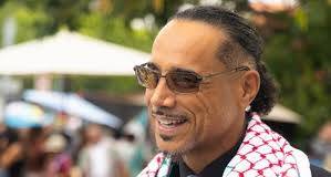 CalCare (universal healthcare) is reintroduced in the California Assembly! Middle aged male of color wearing sunglasses. He has a mustache and a goatee. He is wearing a silver diamond stud in his left ear. Yea is wearing a black shirt with a grey colored tie. His scare is a checkered patten of red, white and green.